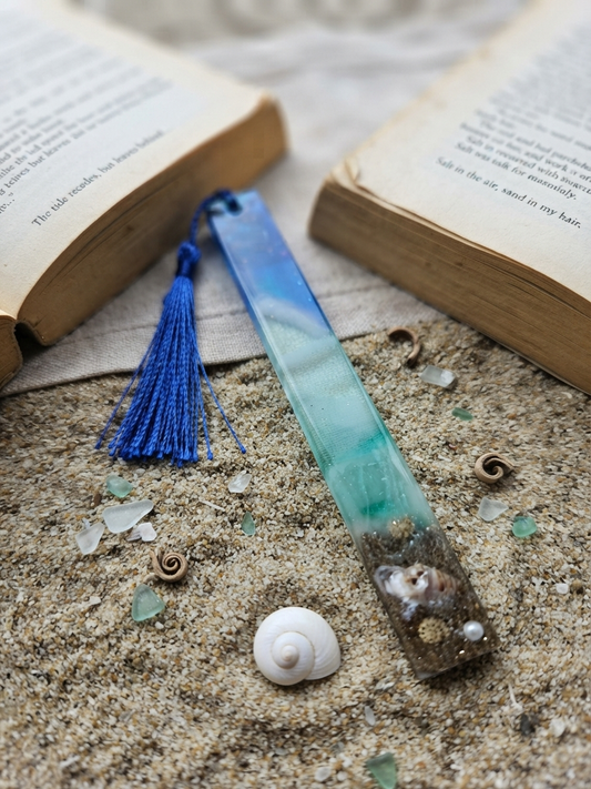 Handmade resin bookmark with blue and teal ocean wave gradient, real sand, seashells, pearl, and cobalt blue tassel, displayed on sand beside an open book