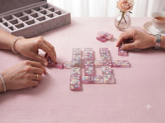 Two people playing handmade cherry blossom resin dominoes on a pink linen table, with grey velvet storage box and rose vase in background