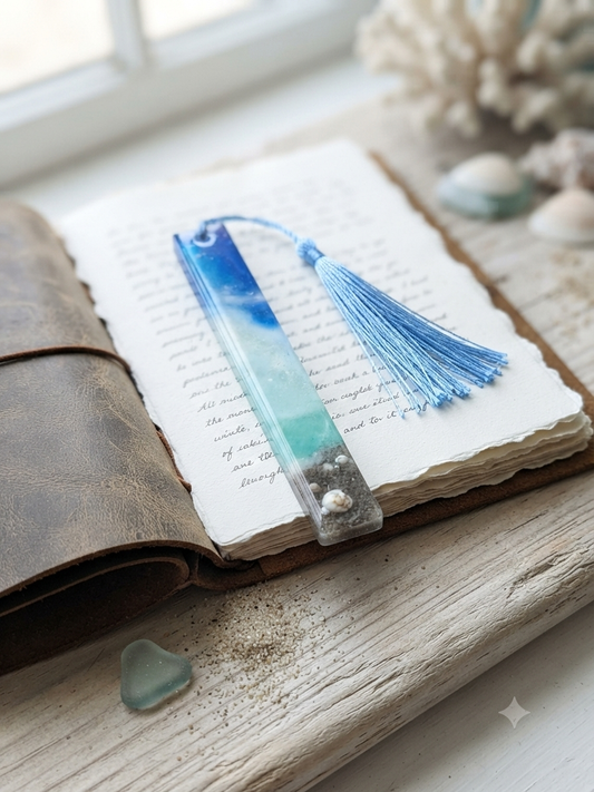Handmade resin bookmark with blue sky and teal ocean gradient, real sand, seashells and pearl at base, with blue tassel, resting on an open journal by a window with coral and sea glass