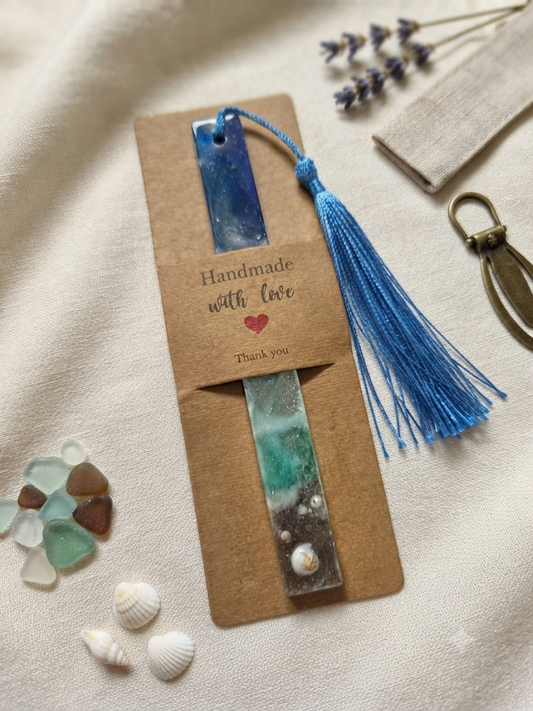 Handmade ocean resin bookmark with blue tassel displayed on a kraft Handmade with Love gift card, surrounded by seashells and sea glass on a linen background