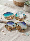 Set of 4 handmade organic-shaped ocean resin coasters with teal and blue waves, real sand, seashells and black pebbles, gold-painted edges, displayed on a driftwood board with a glass of water and seashells