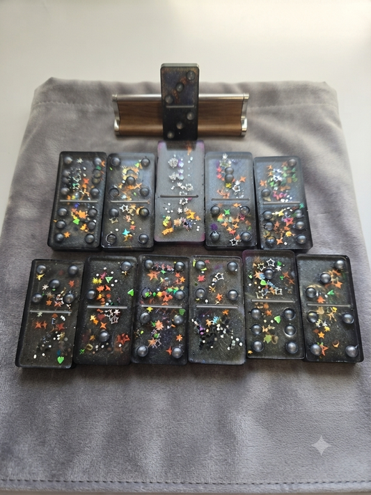 Handmade black resin domino set with holographic multicolour star glitter and grey pearl dots, displayed on a grey velvet pouch with a wooden box holder, arranged in rows