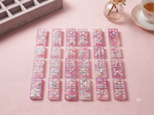 Full set of handmade blush pink resin dominoes with embedded pastel flowers and iridescent glitter, laid out in rows on a pink linen surface with a grey velvet storage box and rose in background